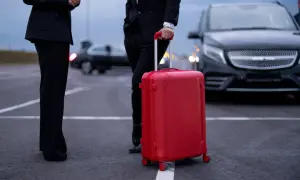 Osoby w garniturach czekają na transport. Czerwona walizka sugeruje, że to delegacja zagraniczna w drodze na ważne spotkanie.