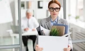 Smutna kobieta z pudełkiem rzeczy osobistych, zastanawia się, kiedy należy się odprawa. W tle siedzi mężczyzna.