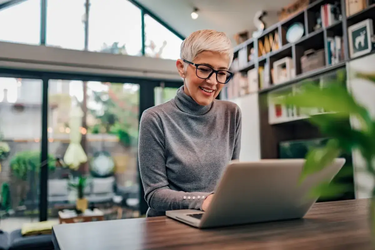 Uśmiechnięta kobieta z krótkimi blond włosami pracuje na laptopie. Może sprawdza swój dodatek do wynagrodzenia.