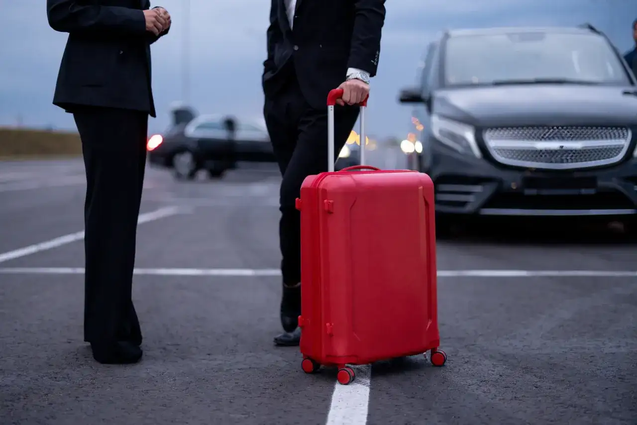 Osoby w garniturach czekają na transport. Czerwona walizka sugeruje, że to delegacja zagraniczna w drodze na ważne spotkanie.