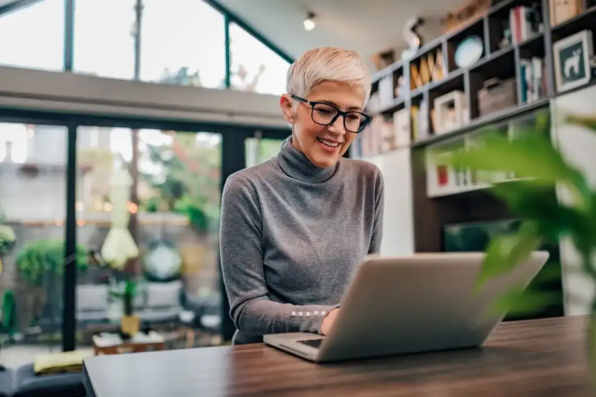 Uśmiechnięta kobieta z krótkimi blond włosami pracuje na laptopie. Może sprawdza swój dodatek do wynagrodzenia.