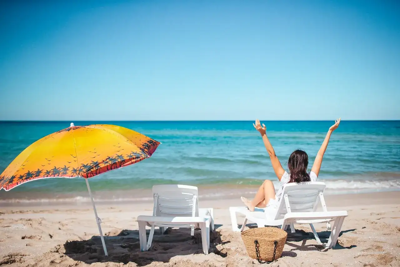 Kobieta cieszy się słońcem na plaży, zastanawiając się, ile dni urlopu jeszcze jej zostało. Obok leżaki i parasol.