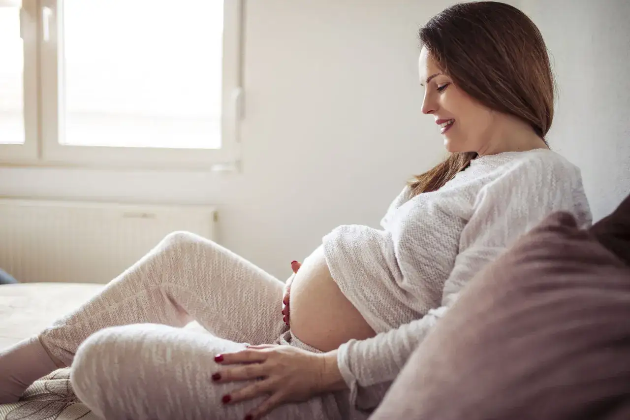 Uśmiechnięta kobieta w 34. tygodniu ciąży, z dłońmi na brzuchu, siedzi na kanapie w przytulnym pokoju.
